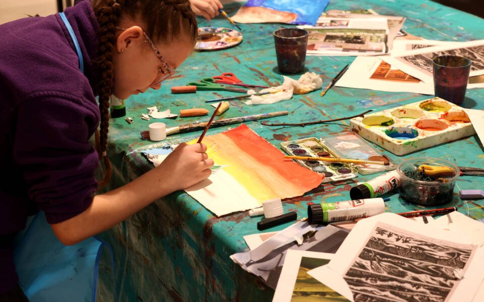 A young girl with glasses paints a sunset landscape using watercolour on paper