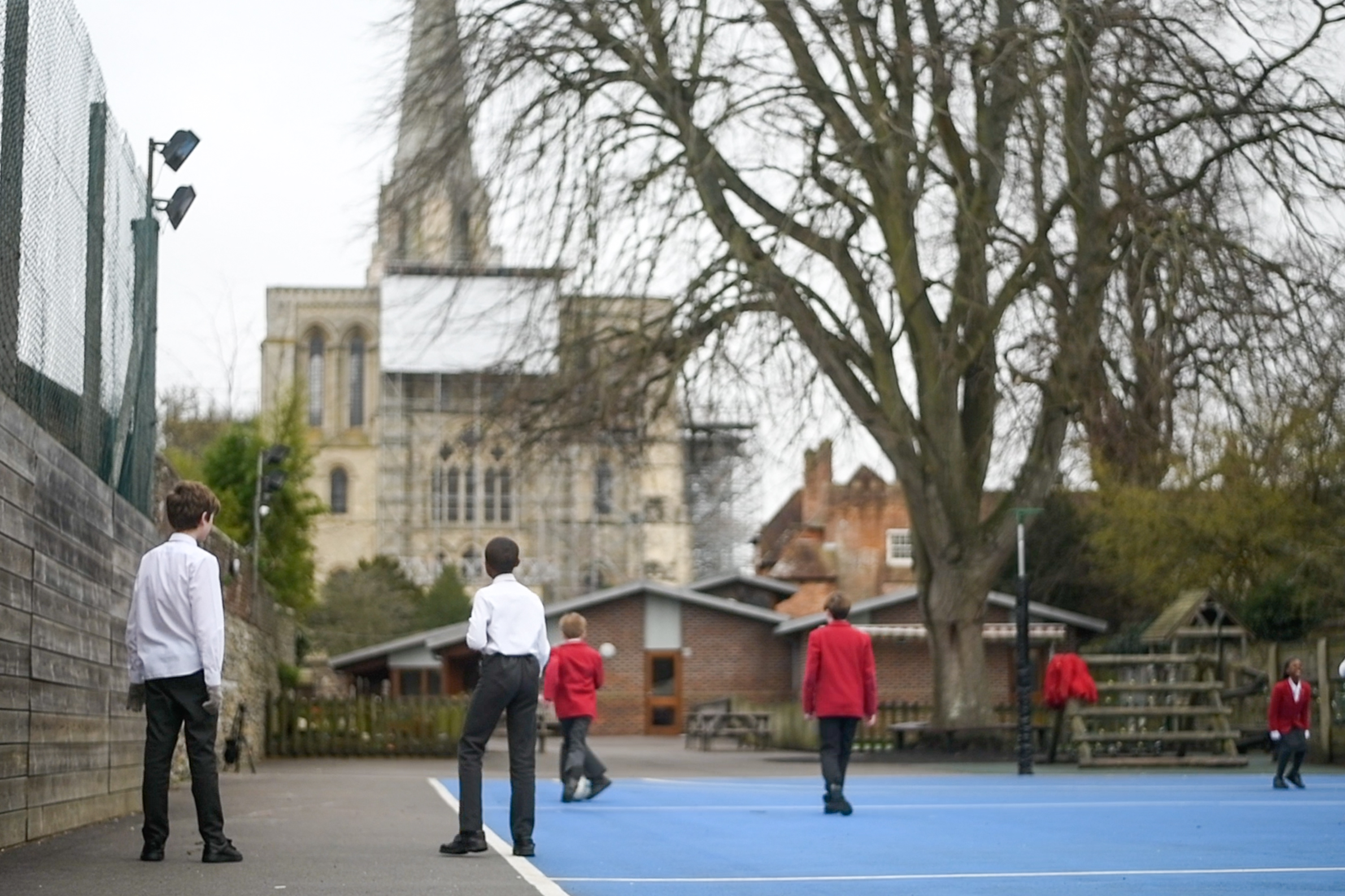 Pupils from the Prebendal School face Chichester's stone Cathedral. The pupils stand on a playground, surrounded by a fence.