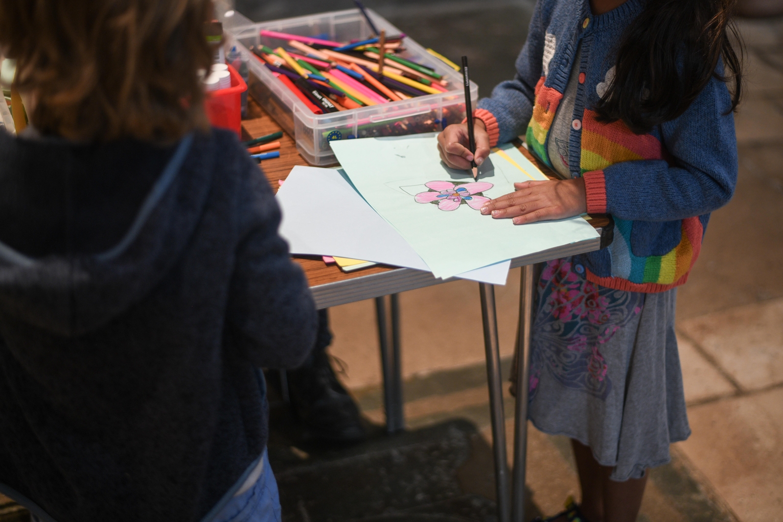 A young girl wearing a rainbow cardigan draws a pink flower on paper using pencils