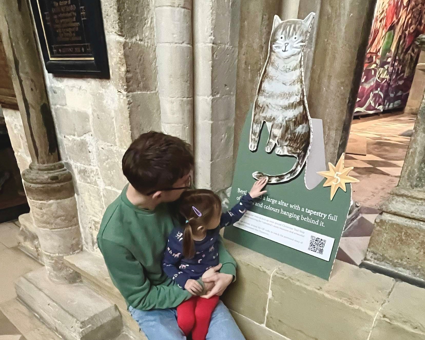 A man holding a child look at a cardboard cut out of a brown and white cat which is sat on stone steps